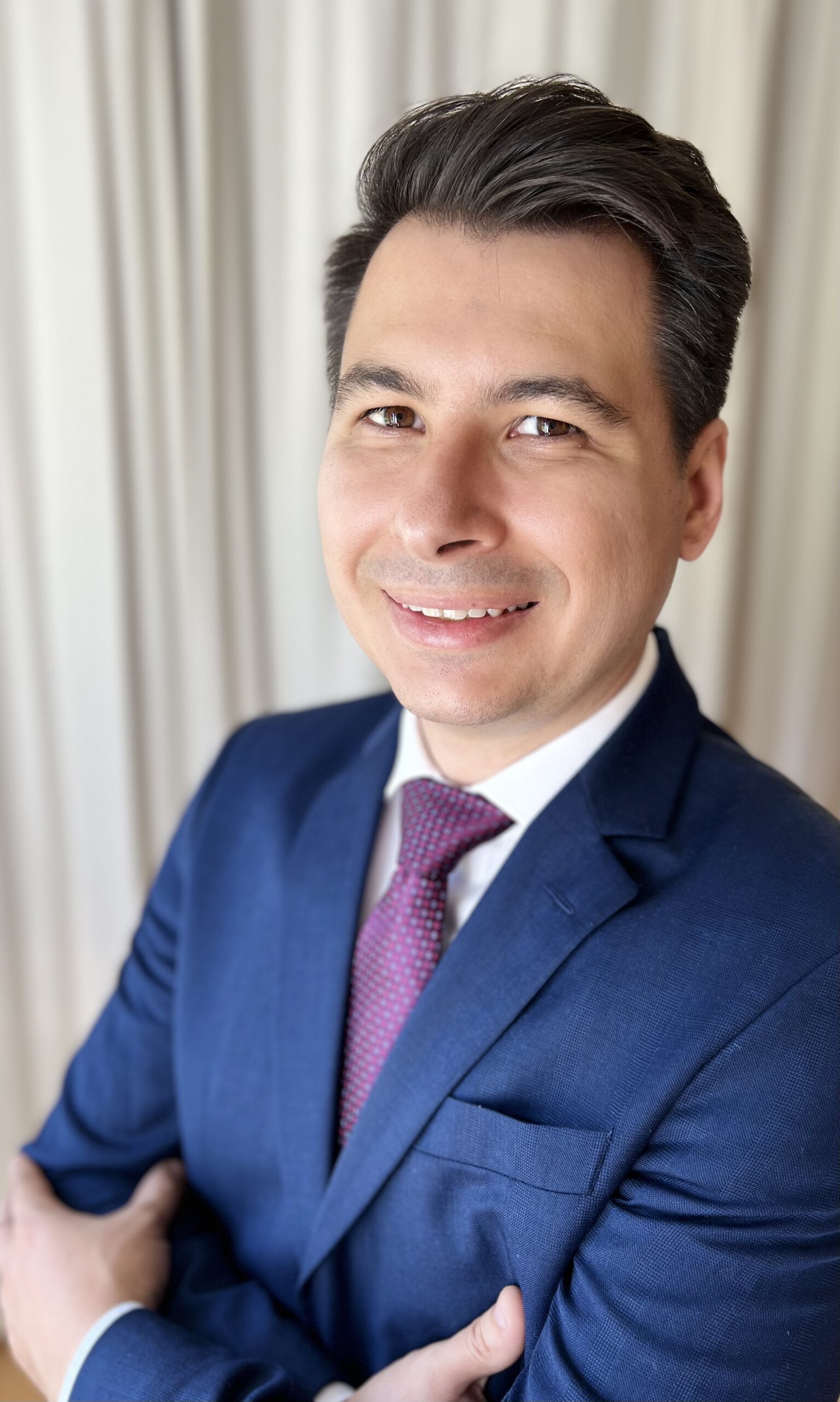 Retrato de Flávio Serpa vestindo um terno azul-marinho, camisa branca e gravata bordô, sorrindo com confiança e braços cruzados. O fundo apresenta cortinas de cores neutras, criando uma aparência profissional e acessível.
